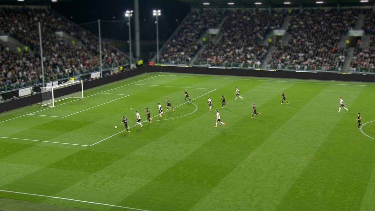 Fernsehbild eines Fußballspiels unter Flutlicht aus der hohen Hauptkamera: grüner Rasen mit Mähstreifen, kleine Spieler im Angriff, volle Ränge im Hintergrund, im Format 16:9 (Symbolbild)