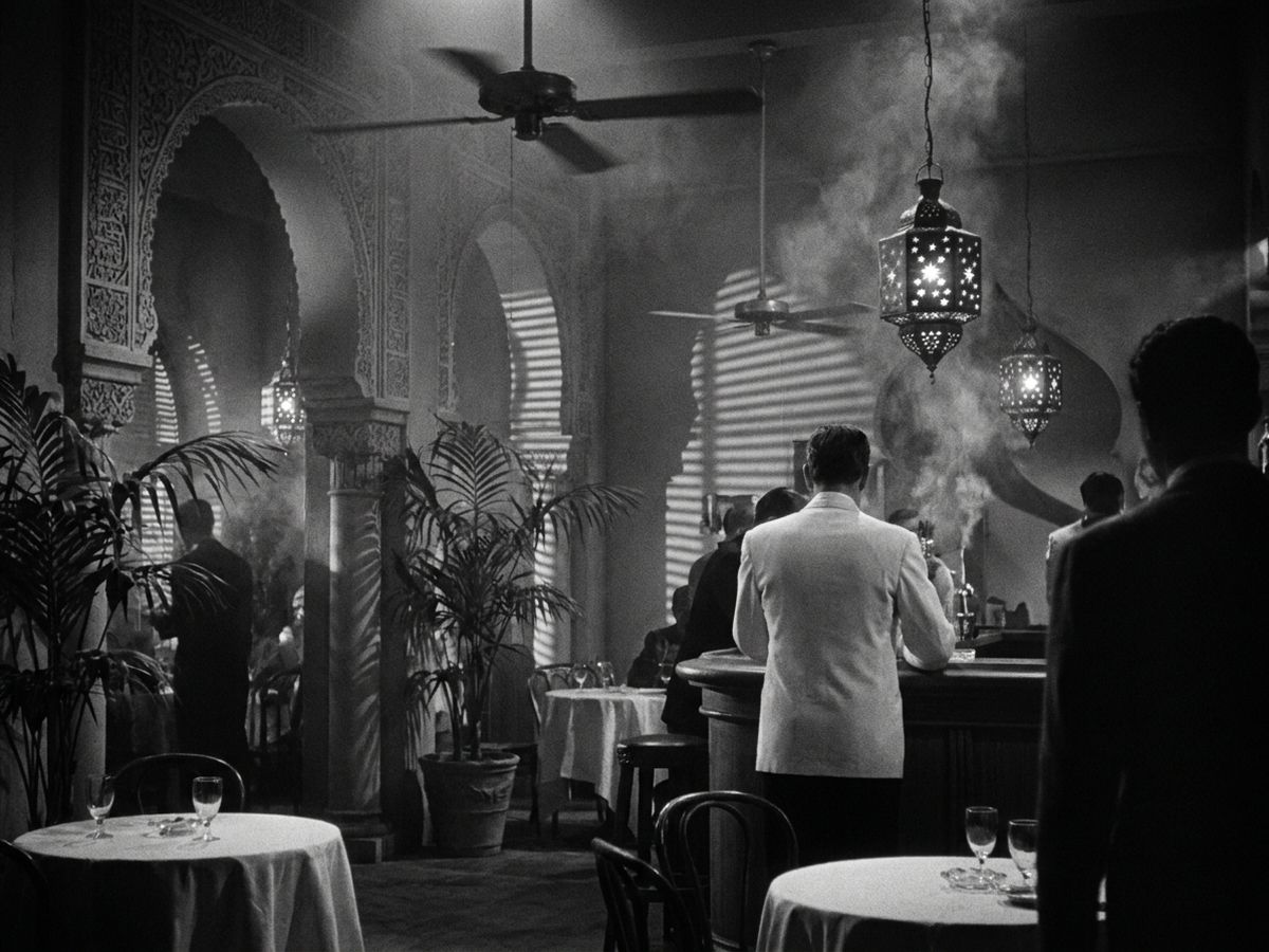 Schwarzweißes Filmbild im Stil der 1940er Jahre: ein verrauchter Nachtclub mit maurischen Bögen, Deckenventilatoren, Palmen und Sternlaternen, ein Mann im weißen Dinnerjackett steht von hinten gesehen an der Bar, im Format 4:3 (Symbolbild)